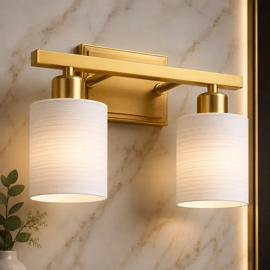 A modern two-light bathroom wall sconce with a brushed gold metal frame and cylindrical white fabric shades, mounted on a beige marble wall.