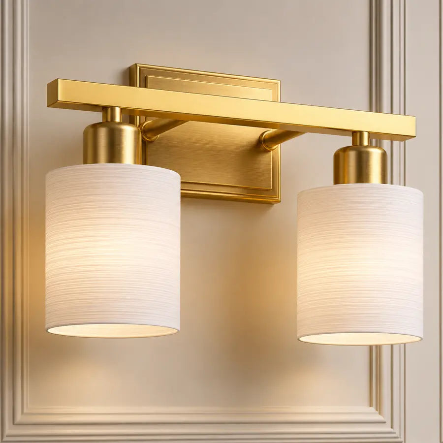 A modern two-light wall sconce with a brushed gold metal frame and cylindrical white fabric shades, mounted on a beige paneled wall.