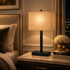 A modern table lamp with a black metal base and a warm beige fabric drum shade, glowing on a marble nightstand in a styled bedroom setting.