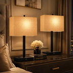 Two matching table lamps with beige fabric shades and black bases sit on a dark wood nightstand in a styled bedroom setting. Decorative books, a small vase of white flowers, pillows, curtains, and wall paneling are visible around them.