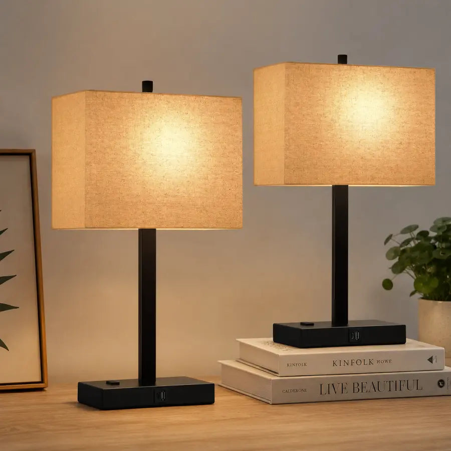 Two modern table lamps with black metal bases and beige fabric shades, shown lit on a wooden surface with books and a small plant in a styled interior setting.