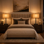 A styled bedroom scene featuring a bed with beige and white bedding, dark brown pillows, a matching upholstered headboard, two matching bedside tables with lit table lamps, and a large abstract painting on the wall.
