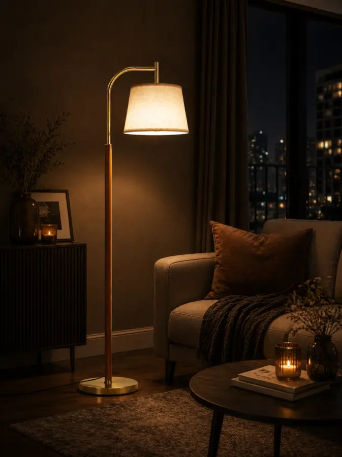 A modern floor lamp with a curved gold-toned metal stand and a white drum shade, glowing warmly in a cozy living room setting next to a sofa at night.