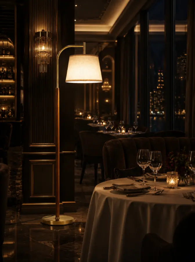 A modern floor lamp with a warm glowing white drum shade and a brushed gold metal frame stands in a dimly lit, upscale restaurant setting with tables set for dining and a city skyline at night visible through the windows.