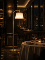 A modern floor lamp with a warm glowing white drum shade and a brushed gold metal frame stands in a dimly lit, upscale restaurant setting with tables set for dining and a city skyline at night visible through the windows.