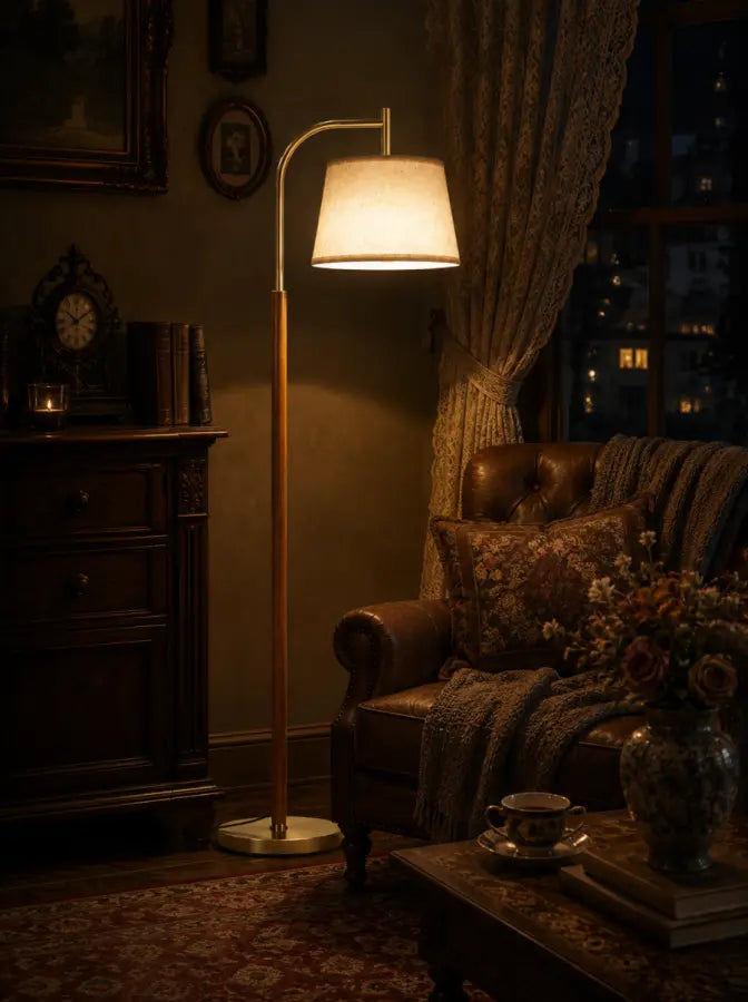 A classic floor lamp with a warm glowing white drum shade and a gold-finished metal pole stands in a cozy, dimly lit living room next to a leather armchair with a patterned throw blanket.