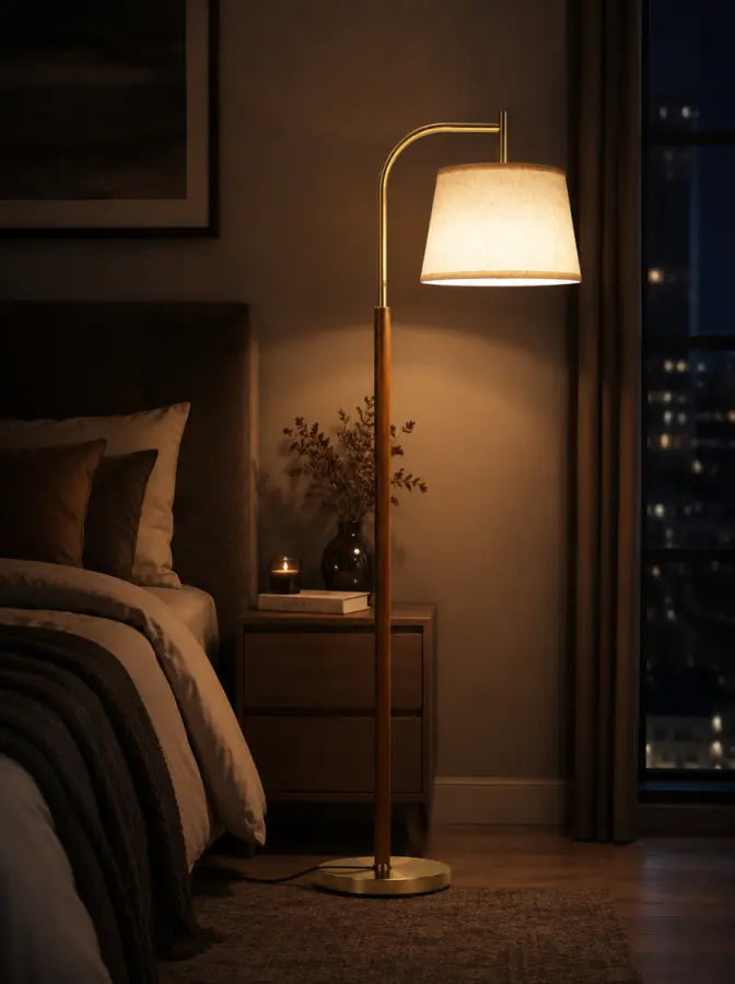 A modern floor lamp with a warm glowing white drum shade, a brushed brass curved arm, and a vertical wooden pole on a round base, shown in a cozy bedroom setting next to a bed.