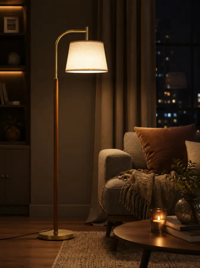 A modern floor lamp with a warm brass-colored metal frame and a white drum shade, illuminated in a cozy living room setting next to a gray armchair with a throw blanket.