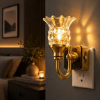 A warm gold wall sconce with a clear textured glass shade, shown lit and mounted on a wall next to a white outlet in a cozy bedroom setting.