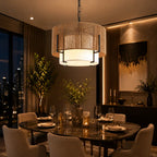 A modern cylindrical pendant light with a woven natural-fiber shade and a glowing white drum diffuser, hanging over a dark round dining table in a luxury, warmly lit contemporary dining room.