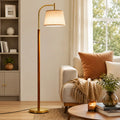 A tall floor lamp with a warm brass-colored metal frame and a beige fabric drum shade stands in a styled living room next to a sofa with cushions and a throw blanket.