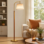 A tall floor lamp with a warm brass-colored metal frame and a beige fabric drum shade stands in a styled living room next to a sofa with cushions and a throw blanket.