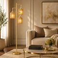A modern floor lamp with a gold-finished metal frame and three warm amber cylindrical shades, styled in a cozy living room next to a beige sofa and round glass coffee table.