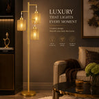 A luxury three-light floor lamp with gold-finished metal frames and warm woven amber glass shades, shown in a styled living room next to a sofa with promotional text on the wall.
