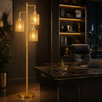 A modern three-light floor lamp with cylindrical woven shades and a brushed gold finish stands in a dark, stylish home office next to a black marble desk and leather chair.