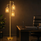 A modern floor lamp with a gold-finished metal frame and three cylindrical woven glass shades, illuminated in a warm light next to a dark desk and office chair.