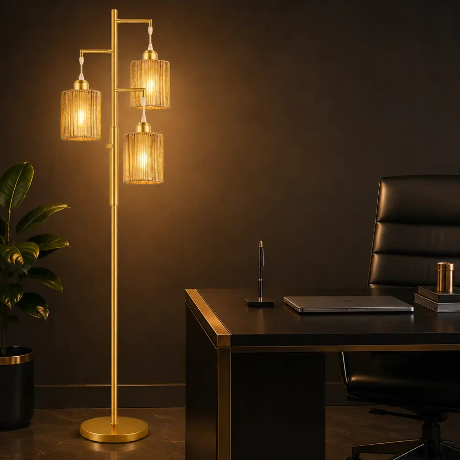 A modern floor lamp with a gold-finished metal frame and three cylindrical woven glass shades, illuminated in a warm light next to a dark desk and office chair.