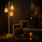A modern three-light floor lamp with cylindrical woven shades and a gold-finished frame, shown lit in a dark, stylishly decorated living room next to a sofa.