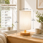 A lit table lamp with a warm white fabric shade and round wooden base sits on a wooden bedside table in a softly styled room. Measurement callouts show the lamp's height, width, and base diameter, with a product dimensions panel on the left listing features like bulb base E26, 6W max, warm white color, 680LM lumens, 120V voltage, and wood + fabric material.