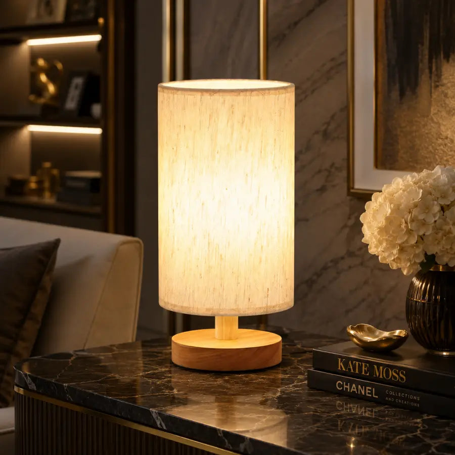 A table lamp with a cylindrical beige textured fabric shade and a round light wood base, illuminated on a dark marble side table in a modern living room setting.