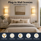 A bedroom scene showing two plug-in wall sconces mounted above nightstands on either side of a bed. The sconces have warm beige fabric shades and are turned on, casting a soft glow.