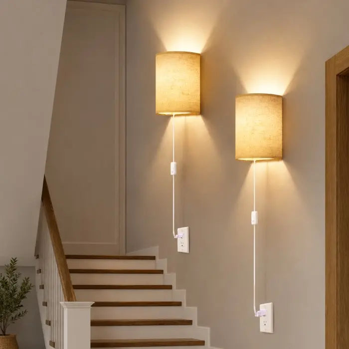 Two cylindrical wall sconces with beige fabric shades and white cords plugged into wall outlets, mounted on a beige wall above a wooden staircase.