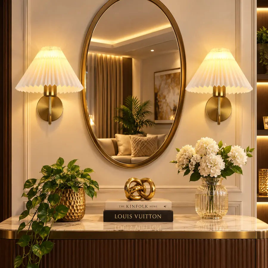 A luxury interior scene featuring a large oval gold-framed mirror on a cream paneled wall, flanked by two matching gold wall sconces with pleated white shades. Below, a marble console table with gold trim holds a potted green plant, a gold decorative object, a Louis Vuitton book, and a glass vase of white flowers.