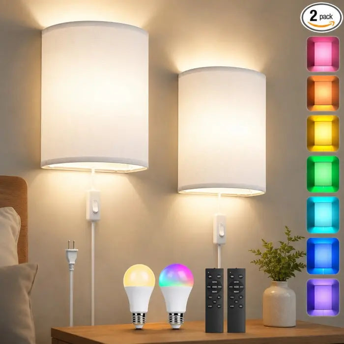 Two white drum wall sconces mounted above a bedside table, each lit with warm white light. Below the lamps are two smart bulb bulbs, one glowing yellow and one multicolor, plus two black remote controls and a small potted plant.