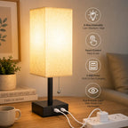 A rectangular table lamp with a beige fabric shade and black base sits on a wooden table in a cozy room setting. The lamp has built-in USB charging ports on the base, with two white chargers plugged in.