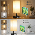 A table lamp with a beige fabric drum shade and black base, shown in a split infographic demonstrating light modes and USB charging capability with two smartphones connected via a white cable.