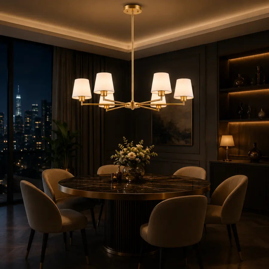 A modern six-light chandelier with white drum shades and a brushed gold finish hangs over a round dark dining table in a dimly lit, upscale dining room with upholstered chairs and a city skyline at night in the background.