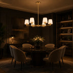 A modern six-light chandelier with white drum shades and a gold-toned frame hangs over a dark round dining table with upholstered chairs in a dimly lit, elegant dining room.