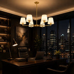 A modern brass six-light chandelier with white drum shades hangs over a dark wood desk in a luxurious, dimly lit home office with a city skyline at night visible through large windows.