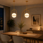 Two modern pendant lights with gold-finished frames and glowing white globe shades hang over a wooden dining table in a warmly lit contemporary dining room.