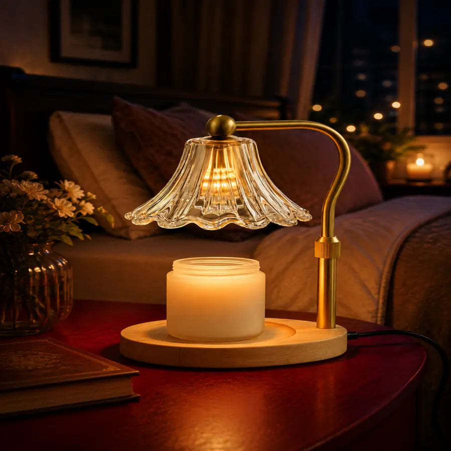 A warm ambient table lamp with a clear scalloped glass shade, gold metal arm, and round wooden base holding a lit white candle, placed on a dark wooden table in a cozy dimly lit living room.