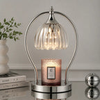 A modern table lamp with a curved silver metal frame and a ribbed clear glass shade, shown lit above a pink textured candle on a reflective surface. The scene is styled with a white vase of greenery and books in the background.