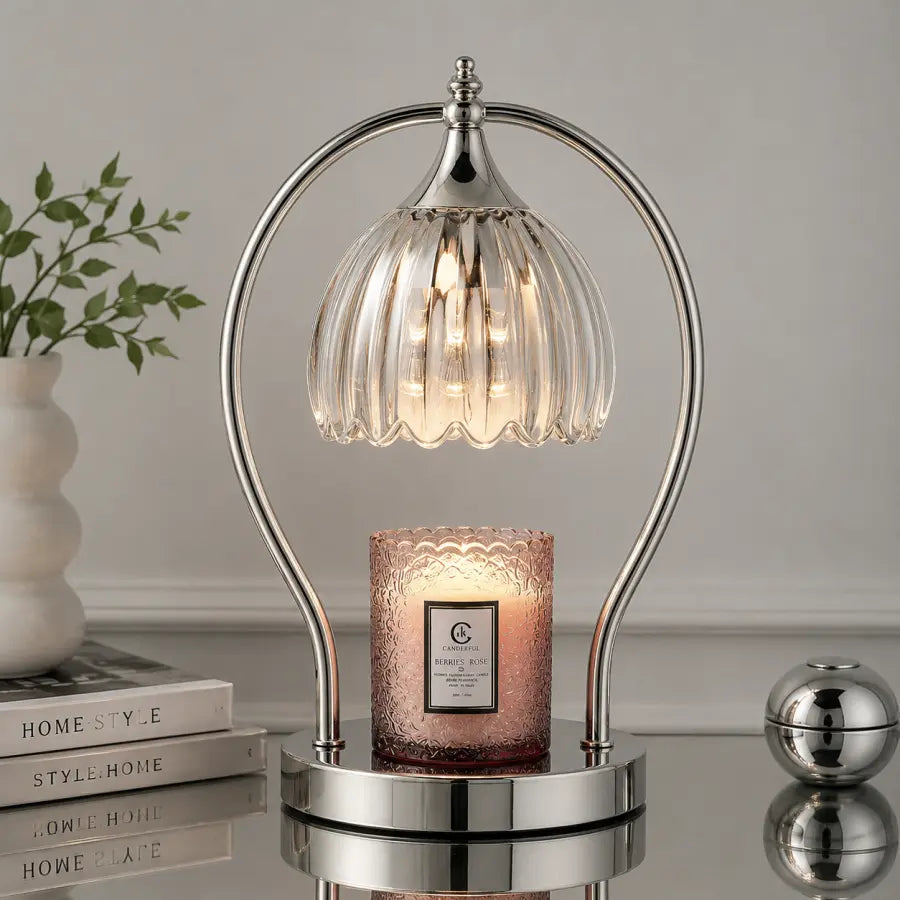 A modern table lamp with a curved silver metal frame and a ribbed clear glass shade, shown lit above a pink textured candle on a reflective surface. The scene is styled with a white vase of greenery and books in the background.