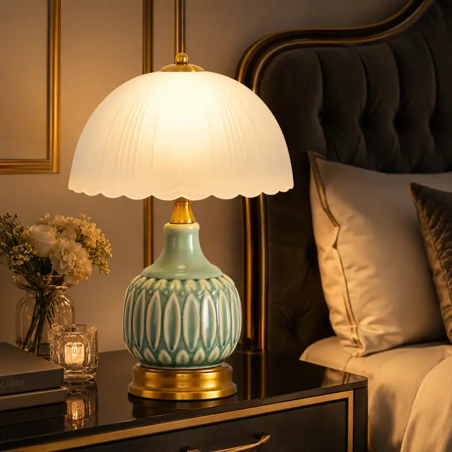 A table lamp with a glowing white pleated dome shade and a light aqua ceramic base with gold accents, placed on a dark bedside table in a bedroom setting.