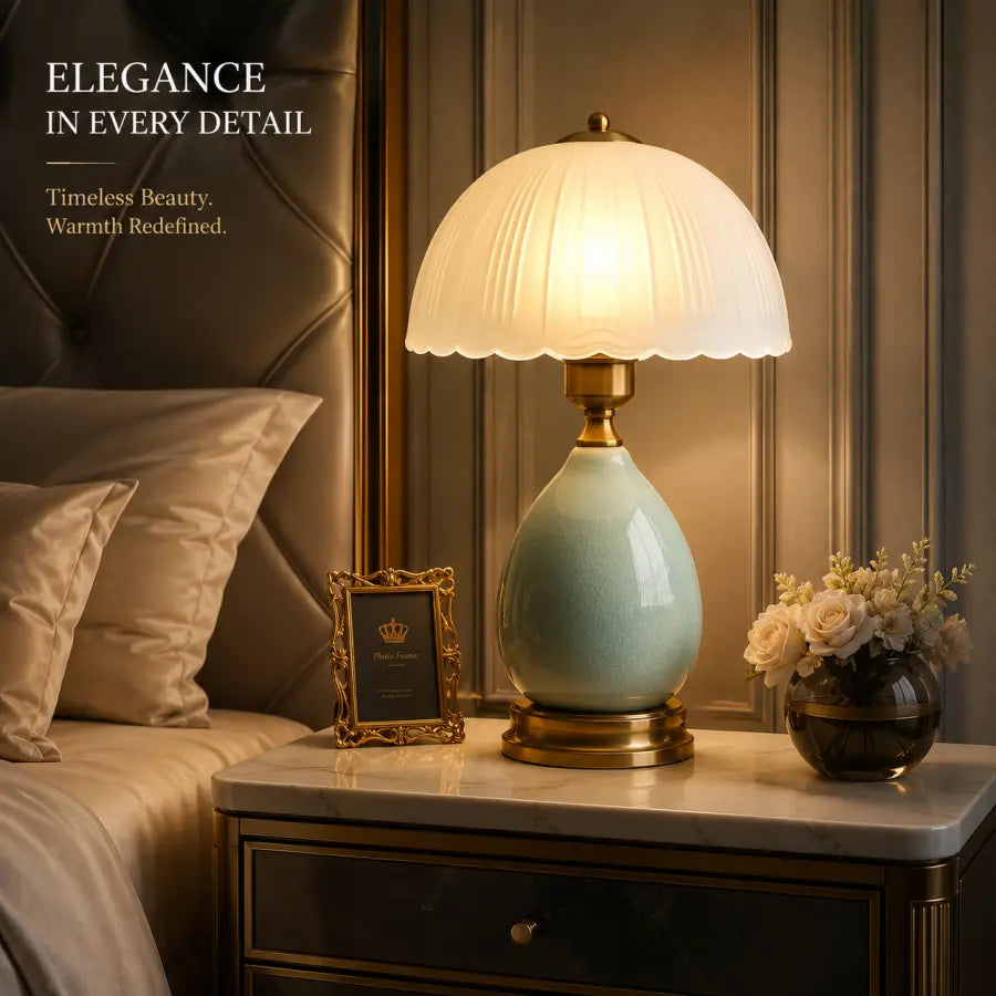 A lit table lamp with a pleated white glass shade and a glossy light blue-green ceramic base on a marble nightstand in a bedroom setting.