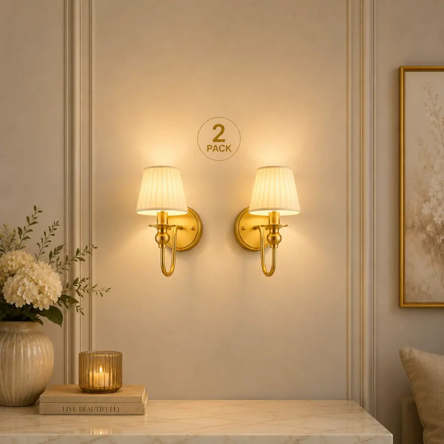 Two matching wall sconces with gold-finished metal mounts and cream pleated shades, shown lit on a beige paneled wall above a marble console. A small '2 PACK' label is visible between the lights.