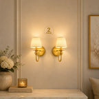 Two matching wall sconces with gold-finished metal mounts and cream pleated shades, shown lit on a beige paneled wall above a marble console. A small '2 PACK' label is visible between the lights.