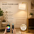 A modern smart floor lamp with a white drum shade and gold-toned metal stand in a living room setting, shown with a smartphone app screen and remote control held by hands at the bottom. Text on the left highlights features like dimmable brightness, color customization, app control, and remote control, with a circular inset showing the adjustable lamp head.