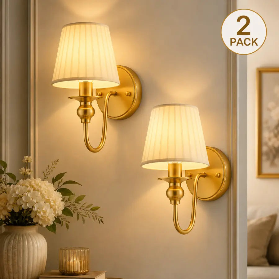 Two gold wall sconces with pleated cream fabric shades mounted on a beige wall, shown in a styled interior with a vase of flowers and a framed picture. A circular '2 PACK' badge appears in the top right.