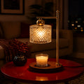 A table lamp with a warm brass-colored metal frame and a faceted clear glass shade glowing with a yellow light, placed on a round red table in a dimly lit living room. A lit candle in a glass holder sits on the table base, with a dark vase and sofa blurred in the background.