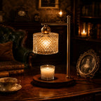 A vintage-style brass table lamp with a faceted crystal drum shade and a curved brass arm, shown lit on a wooden table in a dark, antique room. A small lit candle sits below the lamp, with a teacup and a framed portrait in the background.