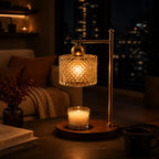 A warm brass table lamp with a faceted crystal drum shade glowing with a yellow light, shown on a wooden coaster on a table in a cozy living room setting. A lit candle in a glass holder sits on the coaster, with a mug and books nearby, and a blurred city skyline at night in the background.