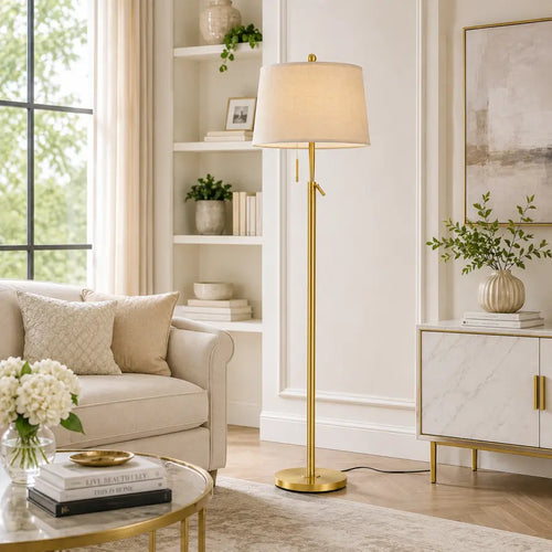 A modern floor lamp with a warm beige fabric drum shade and a polished gold metal stand and base, styled in a bright, elegant living room next to a cream sofa and marble cabinet.
