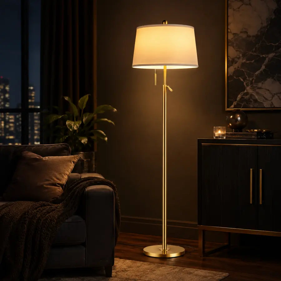 A modern floor lamp with a warm white drum shade and a slim gold metal stand, styled in a cozy, dimly lit living room next to a dark sofa and cabinet.