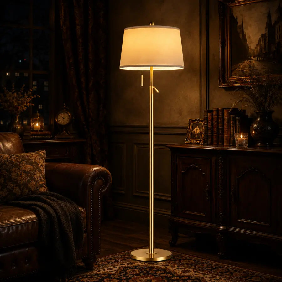 A classic floor lamp with a warm glowing drum shade and a slim gold-finished metal stand, set in a dark, elegant room with a leather armchair and wooden furniture.