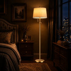 A tall floor lamp with a warm glowing cream fabric shade and a slim brushed metal stand, placed in a dark, elegantly furnished bedroom next to a bed and nightstand.